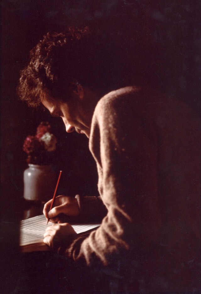 With the only light coming from a window, a man bends over a manuscript with his red pencil. In a stoneware jar on his desk some red flowers. The image is just these elements: man, pencil, flowers, window light.